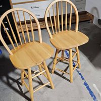Image showing two wooden bar stools side-by-side on concrete floor, highlighting their spindle backs and light wood finish