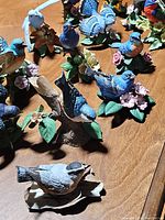 Group photo showing multiple blue porcelain bird figurines on a wooden surface; birds are perched on bases with porcelain leaves and flowers.
