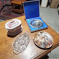 Wide shot of all items on wood surface: gravy boat with cover and base in pink/white, clear glass serving tray, Norman Rockwell decorative plate, and boxed silver-toned commemorative plate with blue satin lining