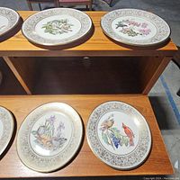 Nine assorted decorative Lenox plates with bird and floral designs, arranged on wood shelf