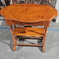 Full view of the antique Victorian wooden table with scalloped top and lower shelf, showing the turned spindle legs and condition of wood.
