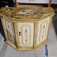 Full view of vintage cream washed wooden cabinet with octagonal shape and ornate door panels. Surface shows wear and tear.