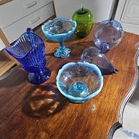 Photo shows six vintage glass pieces in various blue, green, and purple tones displayed on wooden table