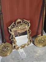 Wide view of the ornate mirror and two brass bed warmers leaning against a red backdrop on the floor.