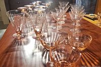 Photo showing arrangement of multiple vintage pink glassware items including stemmed glasses and handled cups placed on a wooden surface.