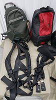 Photo shows two Lowepro Slingshot 100 AW camera bags in green and red/black, along with multiple harness straps laid out on a beige surface, showing details of straps and hardware.