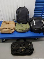 Five laptop bags displayed on a blue bench including beige briefcase, green messenger bag, two black backpacks, and a duffel bag.