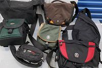 Seven photo and travel bags arranged showing variety including black, brown, green with multiple brand logos visible such as Lowepro, Crumpler, MEC, and ObusForme.