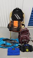 Full lot view showing all six items arranged on and around a chair, including backpacks, messenger bag, hydration pack, and accessories.