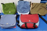 Five Crumpler laptop bags arranged on a surface showing front flap and logo, featuring different colors: green, light blue, tan, dark blue, and red/light blue.