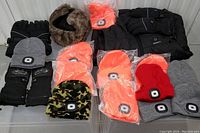 Display of USB rechargeable lighted knit hats in orange, grey, red, and camouflage colors; black insulated gloves; brown faux fur hat; and black insulated vest.
