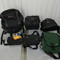Full view of the six camera and video gear bags including two Lowepro LX140 camera bags with tags, MEC backpacks in black and green, Lowepro Orion Mini waist bag, and Lowepro LX160 video photo bag. Shows condition and style of bags.