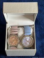 Pair of pink and blue ladies wristwatches neatly placed in a white box on a dark blue background.