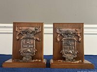 Pair of wooden bookends with bronze University of Toronto coat of arms plaques showing front view, placed upright on blue cloth background.