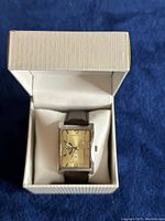 Men's rectangular wristwatch housed in a beige lined box with a gold-tone rectangular dial and brown leather strap.