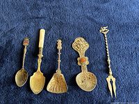 Five collectible sterling silver flatware pieces arranged on blue cloth: three spoons, one triangular scoop with detailed etching, one decorative olive fork.