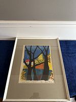 Framed lithograph depicting a colorful, stylized landscape with trees and a large tower, under glass with beige matting, leaning against a wall on a blue carpet.