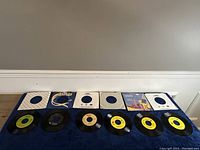 Lot of six 7-inch vinyl records with sleeves and labels visible on some, laid out on blue fabric.