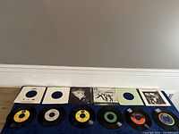 Top down view of all 6 vinyl singles and their sleeves in a row: Queen, 2 Genesis, 2 Madonna, Van Halen