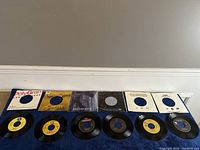 Top view of six 7 inch vinyl records with sleeves laid out on blue cloth, including labels and sleeves showing artists' names and record label details.