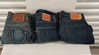 Overview of three pairs of jeans: one True Religion women's and two Levi's 501 men's jeans folded showing the back pockets with brand labels.