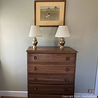 Front view of the wooden 5-drawer dresser topped with two brass lamps and a framed Wyeth print hung on the wall above.