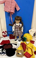 Molly doll with plaid dress and glasses, surrounded by multiple outfits, bags, books, and hats.