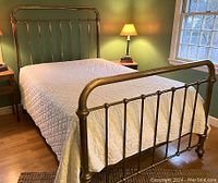 Front angle view of antique brass double bed with quilted white bedding, showing headboard and footboard detail in a bedroom with green walls and wooden floors.