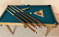 Mini pool table with teal felt surface, four cues on the table with balls in the rack, cue chalk, and felt brush visible.