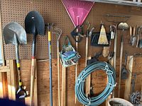Wide view of pegboard wall with various garden tools including shovels, spades, rakes, hose, and hand tools.