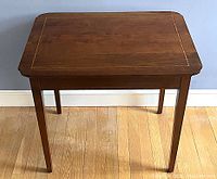 Full view of the solid cherry side table showing shape and leg design on a wooden floor against blue wall.