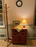 Oak tri-legged coat rack and oak washstand with items on top: ceramic lamp and framed print. Wall clock above the washstand.