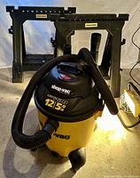Shop Vac in front with two Blue Hawk sawhorses behind, on concrete floor with halogen light casting glow.