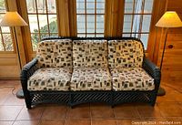 Front view of dark green wicker three-seater couch with patterned cushions, positioned between two floor lamps with shades.