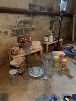 Overview of cake plate, copper molds, glass vases, bowls, and jars arranged on and around small tables on a concrete basement floor.