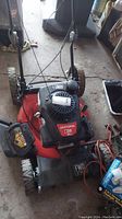 Front angled view of the Craftsman M250 lawn mower showing engine cover, red mower deck, handle, and wheels with visible dirt and items surrounding the mower.