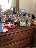 Photo showing five Disney-themed snow globes, two mouse ear crowns, and other figurines on a wooden dresser.