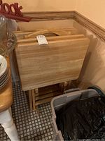 Four wooden folding snack trays stacked upright on matching wooden stand in corner of room.