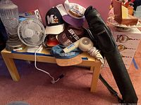 Photo showing a collection of hats with various logos stacked on a small table along with a small white electric fan, a vintage style hair dryer with spiral cord, and a Pro Player pool stick case leaning next to the table.