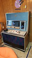 Full front view of Seeburg jukebox showing wood veneer case, blue speaker grille, selection buttons and some records loaded
