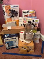 Overall lot photo showing all kitchen appliances mostly in boxes including CucinaPro Pizzelle Maker, Bacon Wave cooker, Maxx Chill cooler, Marcato Atlas pasta maker, Hamilton Beach chocolate fountain, Mr Coffee iced tea maker, and others
