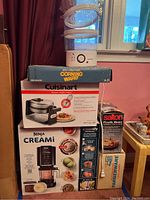 Stacked boxed items showing Corning Ware, Cuisinart waffle maker, Ninja Creami ice cream maker, Salton Pizzelle maker, Farberware colander, and Ovente steamer.