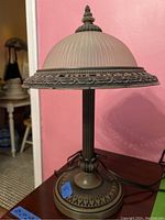 Front angled view of the heavy metal lamp with ribbed glass shade and decorative metal trim on a wooden surface with a pink wall background