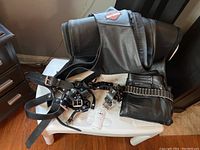 Photo showing folded black leather vest with Harley Davidson patch on back, along with straps, belt, and folded leather pants