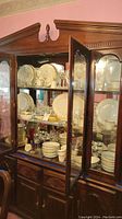 Wooden china cabinet with glass doors displaying white Lenox china with gold trim, assorted glassware, and serving pieces.
