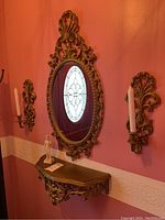 Ornate plastic oval mirror with carved details and crest, mounted over matching carved shelf, with a small David statue on it, flanked by two matching wall sconces holding white taper candles, all against a pink wall.