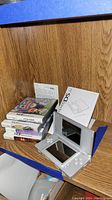 Photo of Nintendo DS Lite console, instruction manuals, and 5 game cases stacked beside it on a wooden surface.