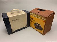 Two vintage projectors side by side, one in a hard carrying case and the other in its original cardboard box.