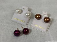 Three pairs of pearl stud earrings displayed on cloth. White, brown and dark purple pearls, marked 14K gold.