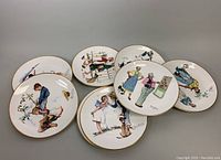 Eight vintage Norman Rockwell collectible porcelain plates with various colorful illustrations and gold rims arranged on a surface.
